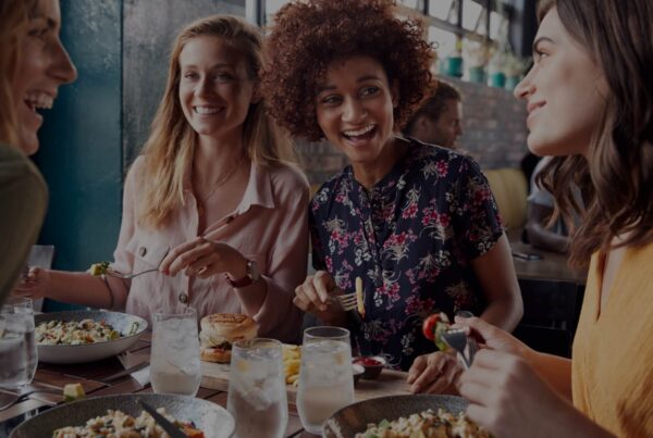 Four women sit together at a restaurant table, smiling and laughing while enjoying salads and sandwiches. With plates of food and glasses of water, they create a cheerful, social vibe—perfect for exploring the New LAX Shops & Eateries.