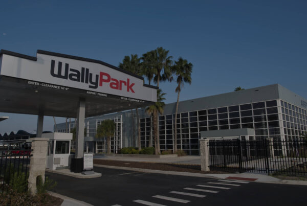 A WallyPark parking facility with a large entrance canopy, palm trees, and a modern glass building under a clear blue sky. The sign reads WallyPark with entry and clearance information.