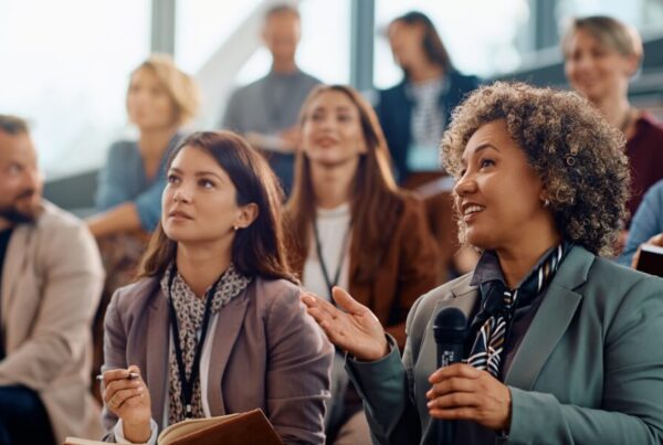 A diverse group sits in an auditorium; a woman in front, holding a microphone, speaks while others listen and take notes at one of the Most Popular Conventions of 2024—a professional, collaborative setting.