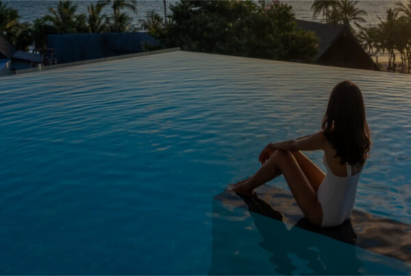 A woman in a white swimsuit sits at the edge of an infinity pool, embracing the quiet travel trend as she gazes at the sunset over the ocean, with palm trees and buildings silhouetted in the background.