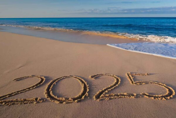 The number 2025 is written in large letters in the sand on a beach, with gentle ocean waves approaching and a blue sky in the background.