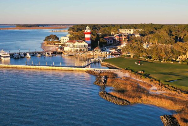 Aerial view of a marina with boats, a red-and-white striped lighthouse, buildings, and a golf course—perfect for a family spring break getaway. The coastline, dotted with trees and water, shines under a clear sky at sunset.