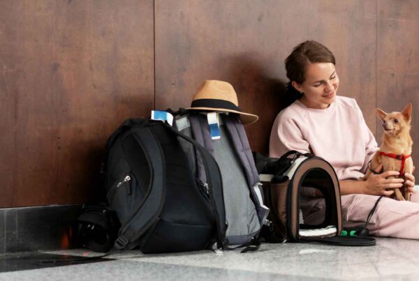 A woman in pink sits on the floor with her legs outstretched, holding a small brown dog—surrounded by backpacks, a hat, and a pet carrier—waiting in an airport during family spring break travels.