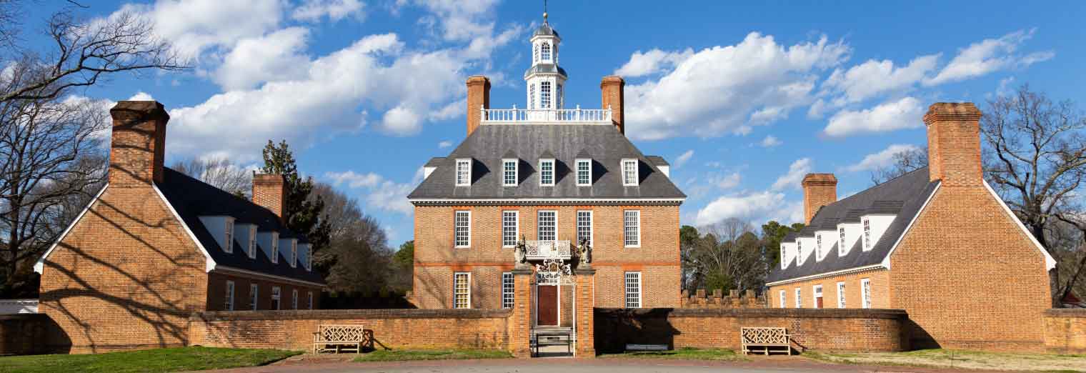A large historic brick building with a central cupola, flanked by two smaller brick wings, sits under a blue sky with scattered clouds—an inviting spot for a family spring break—surrounded by a low brick wall.
