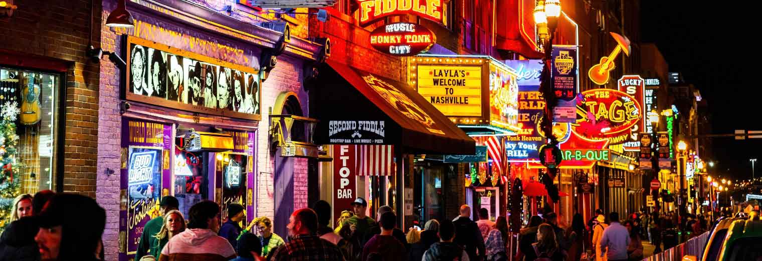 A lively street scene at night in Nashville, Tennessee, with colorful neon signs for bars and music venues, and a crowd of people—including families on spring break—walking along the sidewalk.