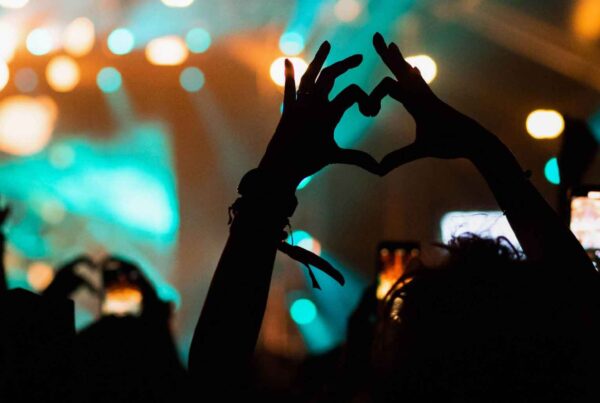 Silhouetted crowd at a concert with colorful stage lights in the background; one person forms a heart shape with their hands while others, enjoying family spring break, hold up phones to capture the moment.