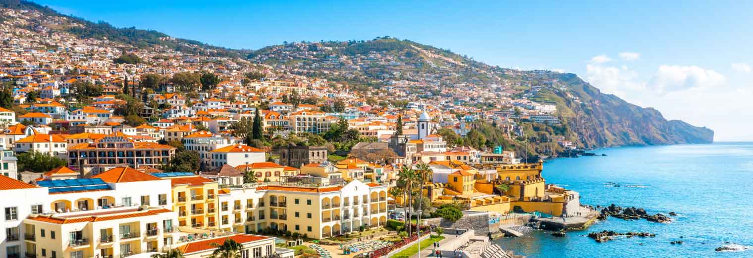 Colorful buildings with terracotta roofs line the coast of a hilly island, perfect for a family spring break, with mountains rising behind and the blue ocean stretching out under a clear, sunny sky.