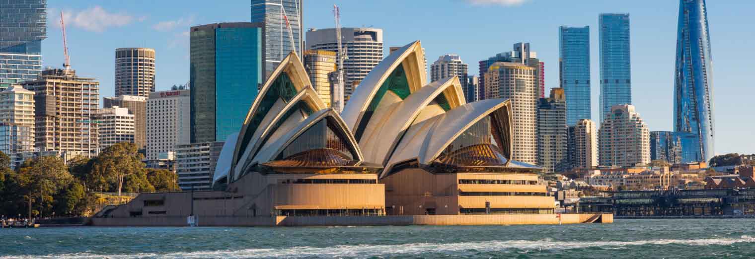 The Sydney Opera House with its distinctive sail-like roofs sits by the water, making it a perfect family spring break spot, surrounded by modern skyscrapers and city buildings under a clear blue sky.