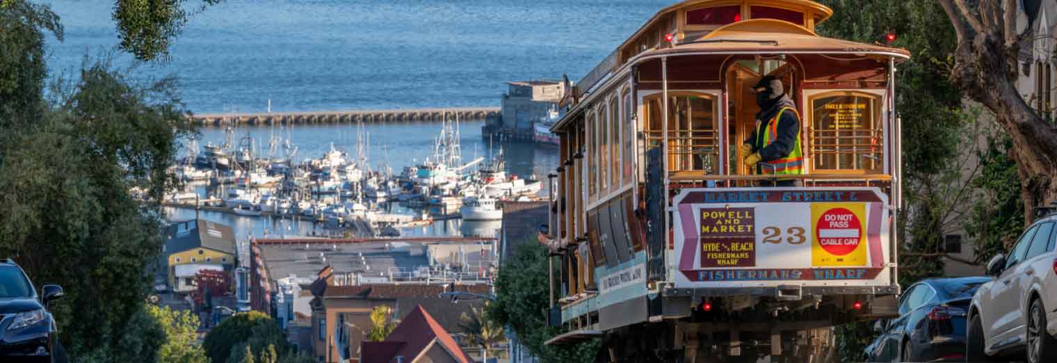 A San Francisco cable car travels downhill toward a marina filled with boats, with the bay and city buildings in the background—a perfect family spring break scene, with trees and parked cars lining the steep street.