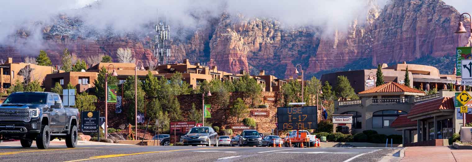 A scenic street in Sedona, Arizona, perfect for a family spring break, with cars driving by, red rock cliffs in the background, low clouds, and several buildings, shops, and road signs lining the road.