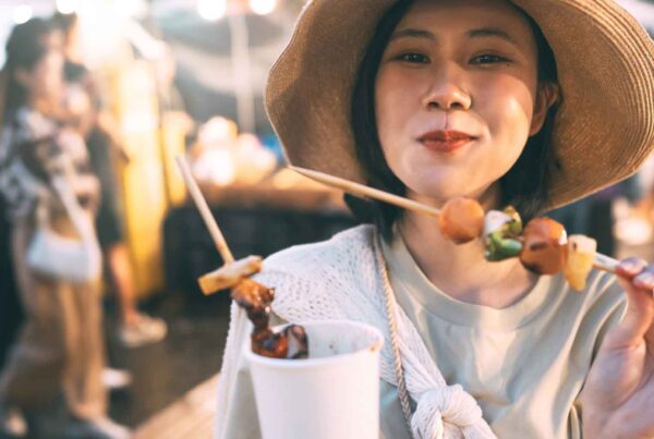 A woman wearing a wide-brimmed hat smiles while holding skewers of grilled food and a cup at an outdoor night market—an experience found in great cities for fan favorite foods, with blurred people and stalls bustling in the background.
