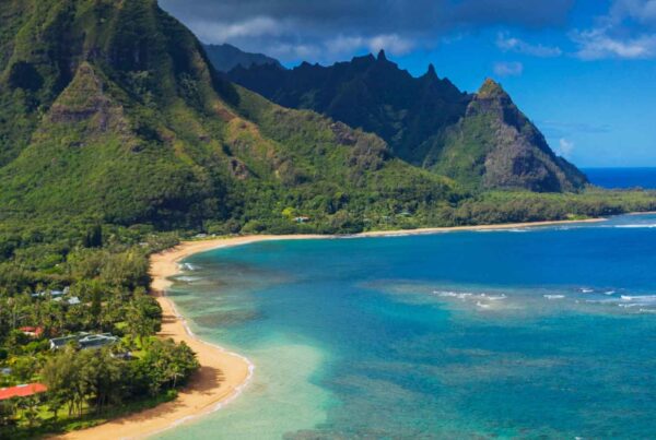 Aerial view of a lush green mountain range beside a sandy beach and clear blue ocean, with gentle waves and scattered houses nestled among tropical trees—one of the top cheap fall travel destinations.