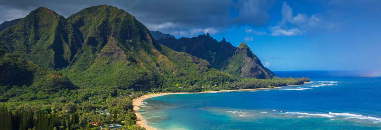 wallypark-airport-parking-fall-deals-inset1 A lush green mountain range rises beside a sandy beach and clear blue ocean under a partly cloudy sky—one of the stunning cheap fall travel destinations with waves gently rolling into the shore.