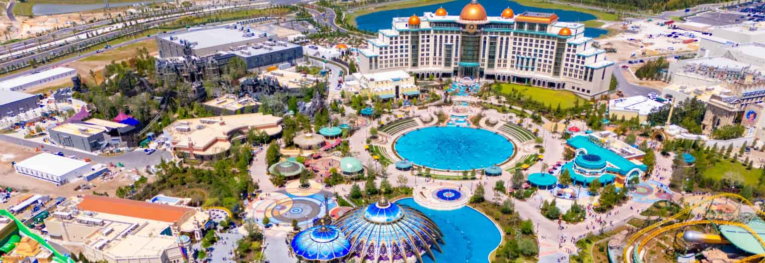 wallypark-airport-parking-fall-deals-inset2 Aerial view of a large, colorful amusement park—one of the top cheap fall travel destinations—with multiple pools, dome-shaped structures, and a grand hotel surrounded by landscaped gardens and various attractions.