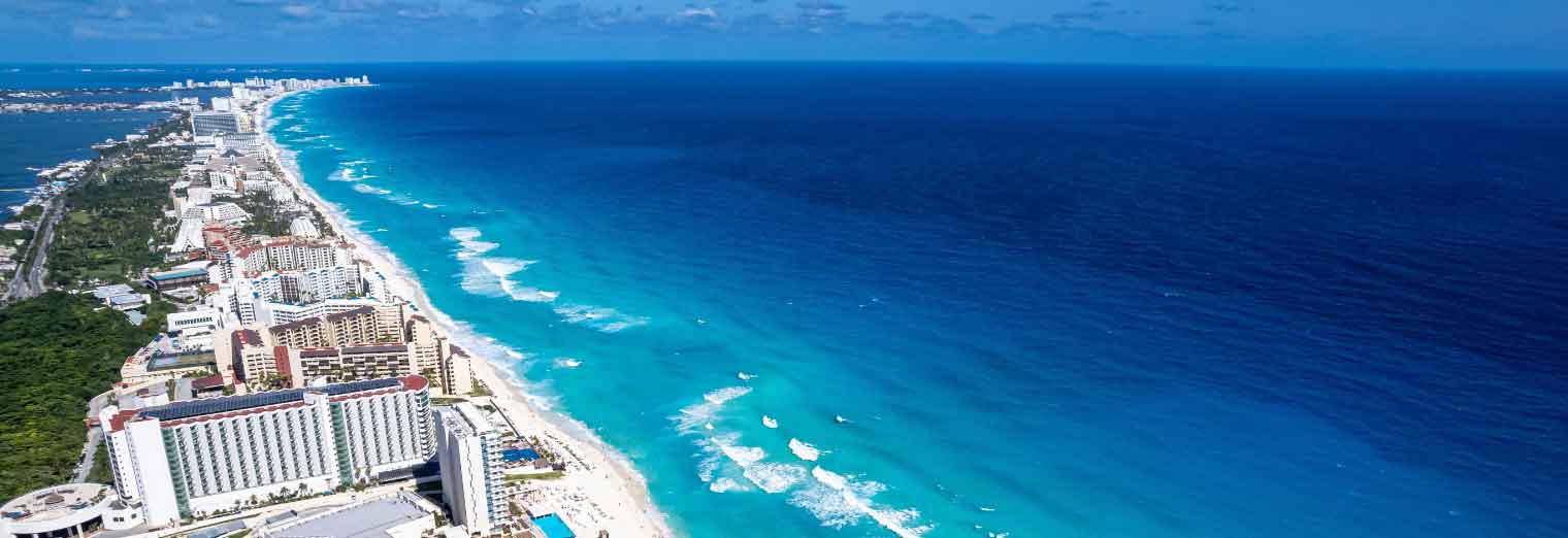 wallypark-airport-parking-fall-deals-inset4 Aerial view of a long stretch of hotels and resorts lining a white sandy beach—one of the top cheap fall travel destinations—with turquoise ocean waves on one side and a lagoon on the other under a sunny blue sky.