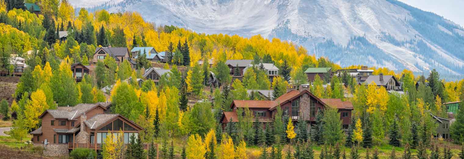 wallypark-airport-parking-fall-deals-inset6 A cluster of wooden houses nestled among green and yellow autumn trees at the base of a tall, rugged mountain—an idyllic scene perfect for those seeking cheap fall travel destinations.