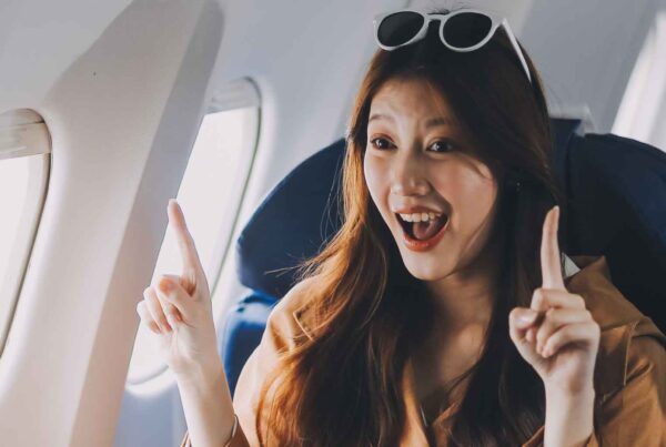 A young woman with long hair, wearing sunglasses on her head, sits by an airplane window and smiles excitedly while pointing upward with both index fingers, ready to explore cheap fall travel destinations.