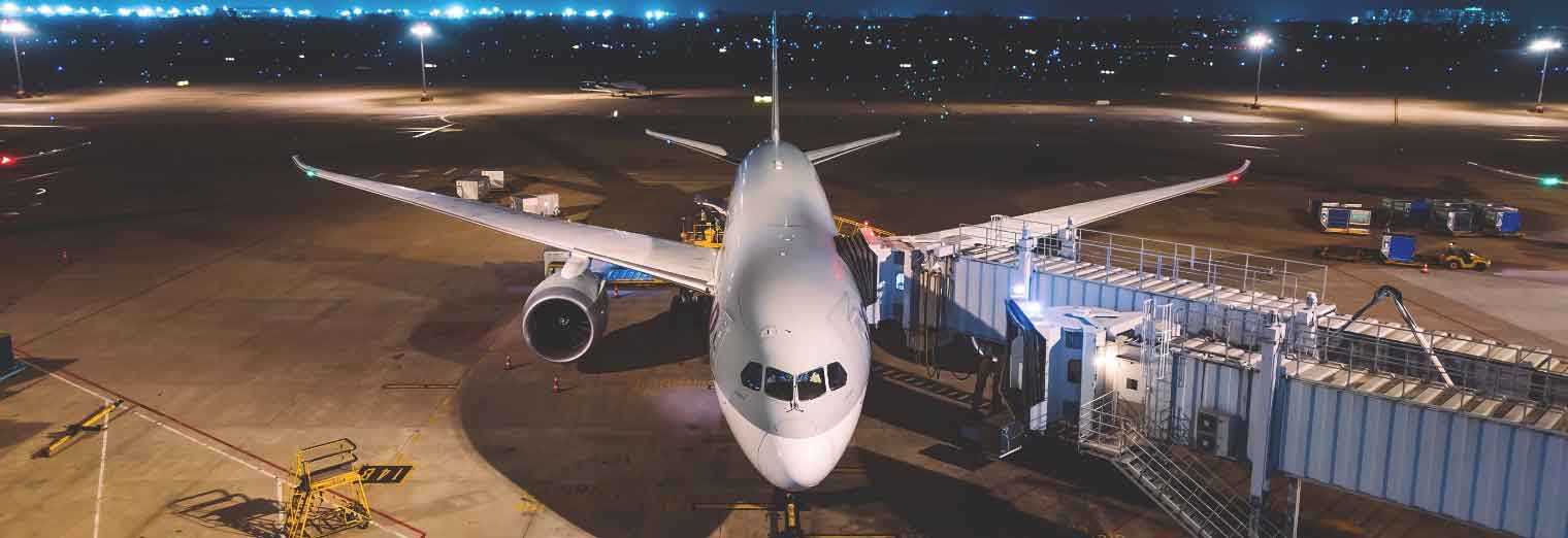 A large passenger airplane is parked at an airport gate at night, connected to a jet bridge, with bright lights illuminating the runway—ready to whisk travelers away to cheap fall travel destinations as city lights twinkle in the distance.
