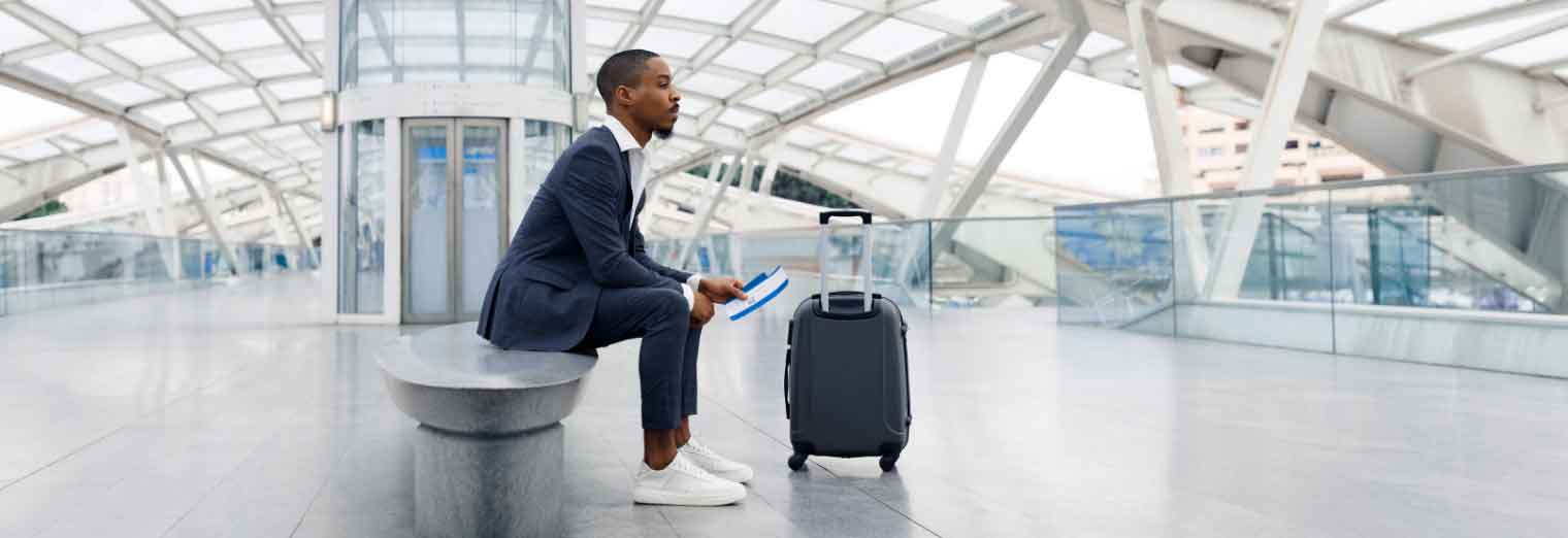 A man in a suit sits on a bench in a modern, glass-covered terminal, holding a tablet and boarding pass—ready to explore cheap fall travel destinations. A rolling suitcase stands beside him.