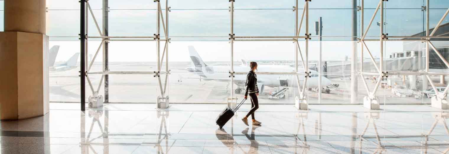 A person walks through an airport terminal, pulling a wheeled suitcase, excited to explore cheap fall travel destinations. Large windows reveal airplanes parked outside on the tarmac under a bright sky.