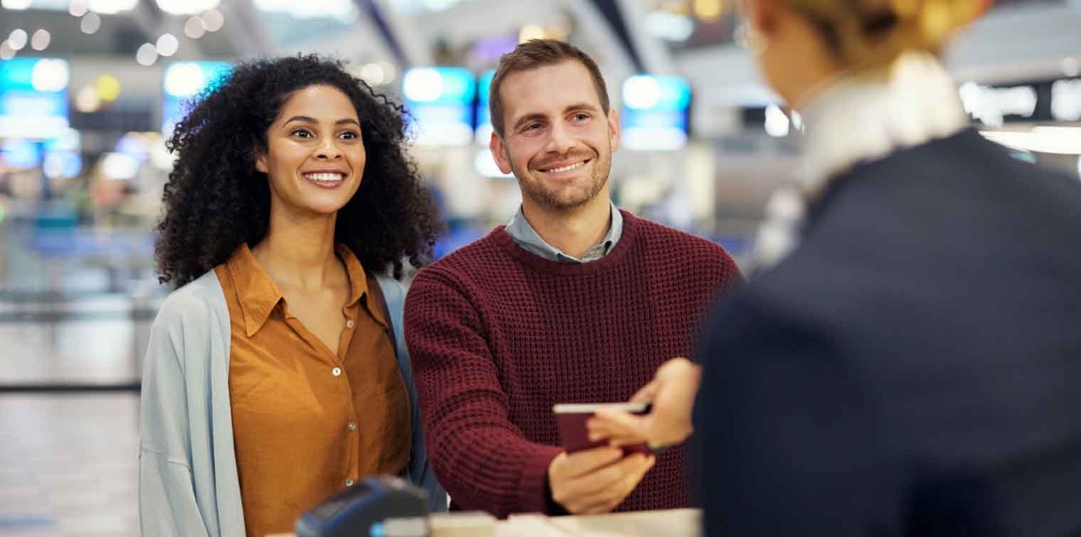 A smiling couple stands at an airport check-in counter, handing passports to an airline staff member. The bright, modern terminal hints at their excitement as they embark on cheap fall travel destinations.