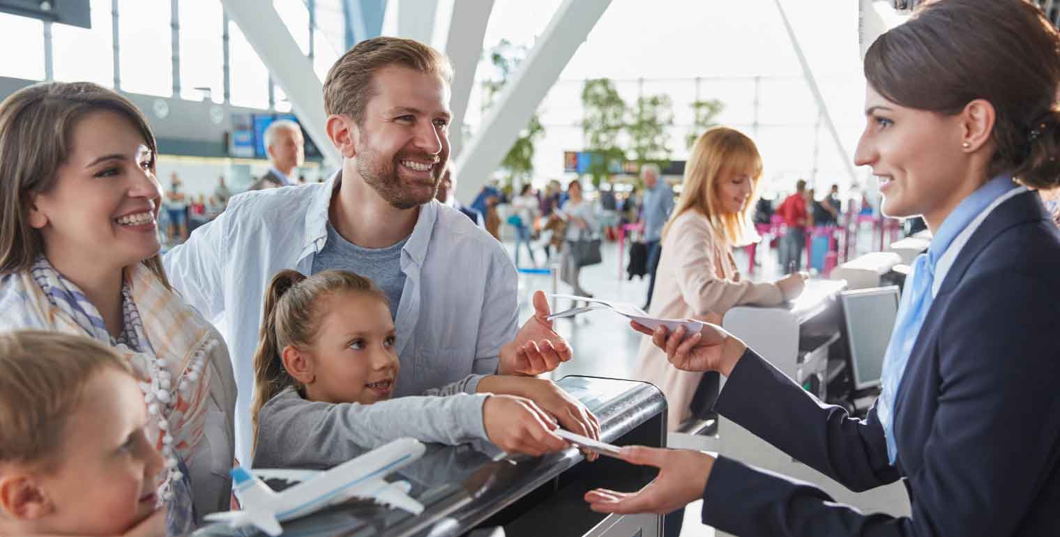 A smiling family with two children checks in at an airport counter, speaking with a friendly airline staff member about their cheap fall travel destinations. The airport is busy with travelers in the background.