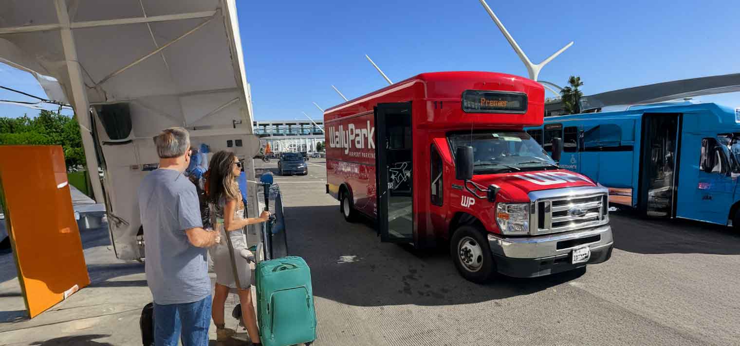 People with luggage wait at a bus stop as a red LAX FlyAway shuttle approaches, ready to whisk travelers to cheap fall travel destinations, while another blue shuttle sits parked nearby under a clear, sunny sky.