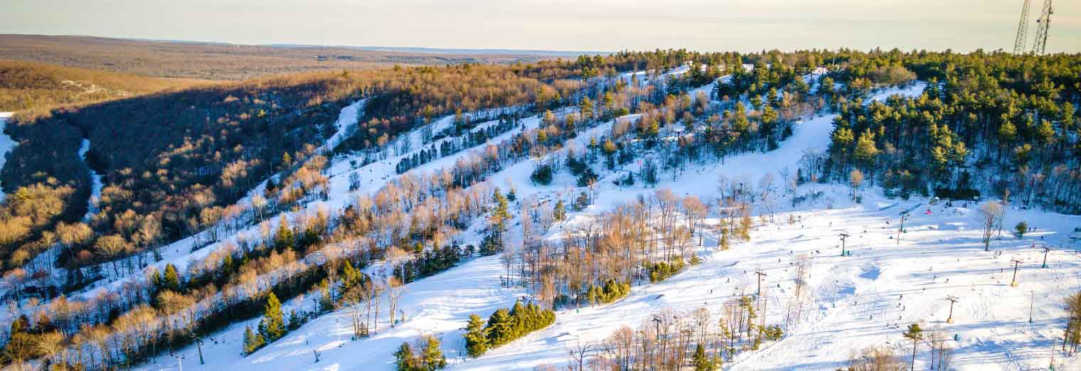Aerial view of a snow-covered ski slope with scattered trees, ski lifts, and skiers. Forested hills surround the area under a clear sky, with sunlight casting long shadows across the landscape.