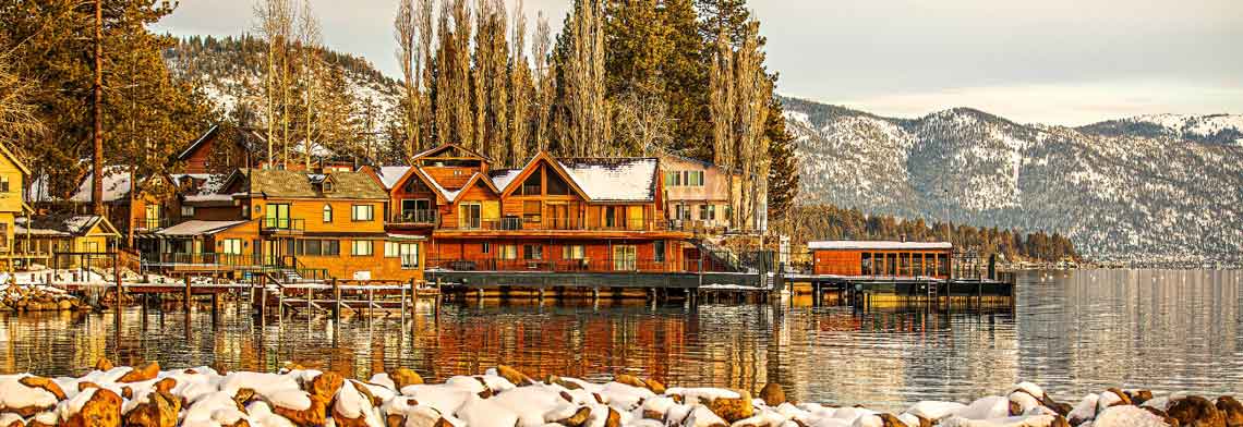 Lakeside cabins with snow-covered roofs and docks sit beside calm water, surrounded by pine trees and snow-dusted mountains in the background, under a cloudy sky.
