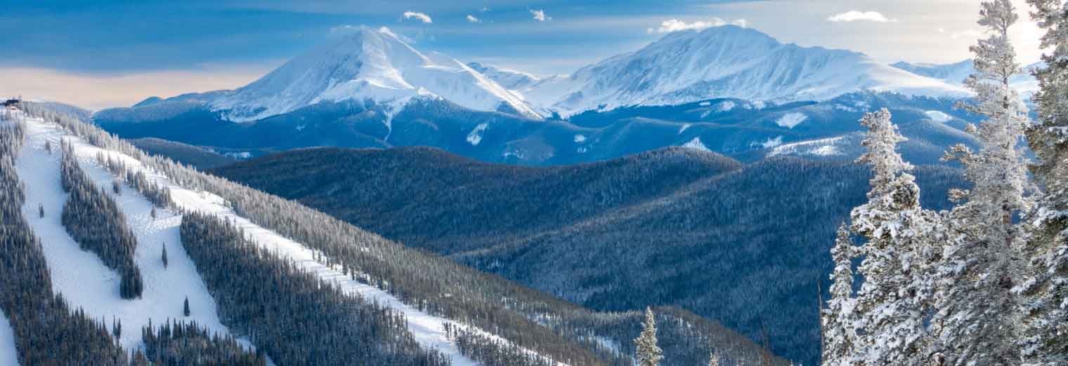 Snow-covered mountains and trees under a clear blue sky; ski slopes with tracks visible on the left, with tall peaks and forested hills stretching into the distance.