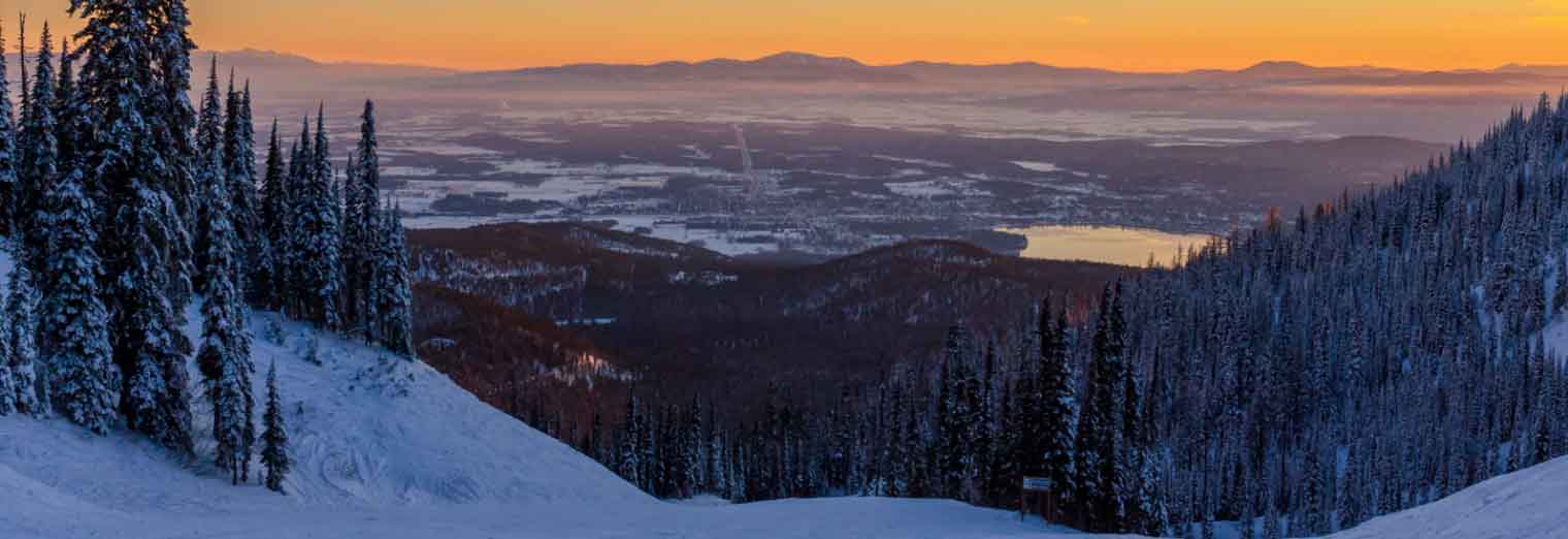 Snow-covered mountain slopes lined with pine trees overlook a valley and frozen lake at sunset, with orange, pink, and purple hues in the sky and distant mountains on the horizon.