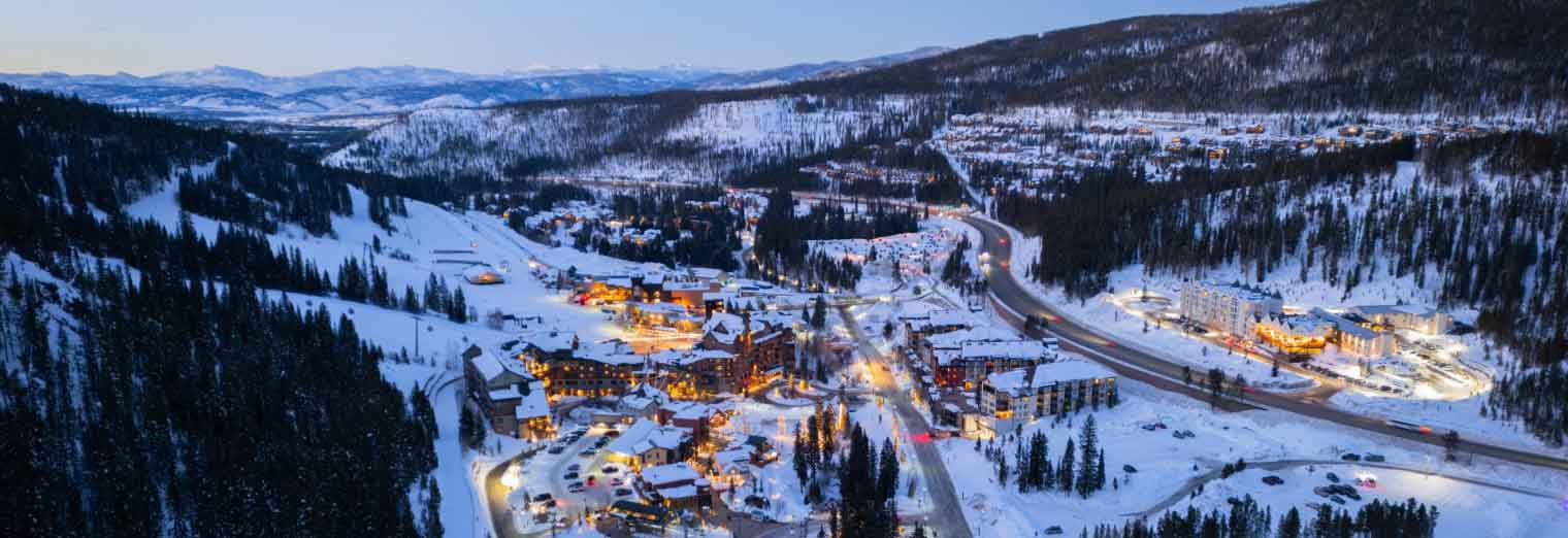 Aerial view of a snow-covered mountain village at dusk, with warmly lit buildings nestled among pine trees, winding roads, and mountains in the background.
