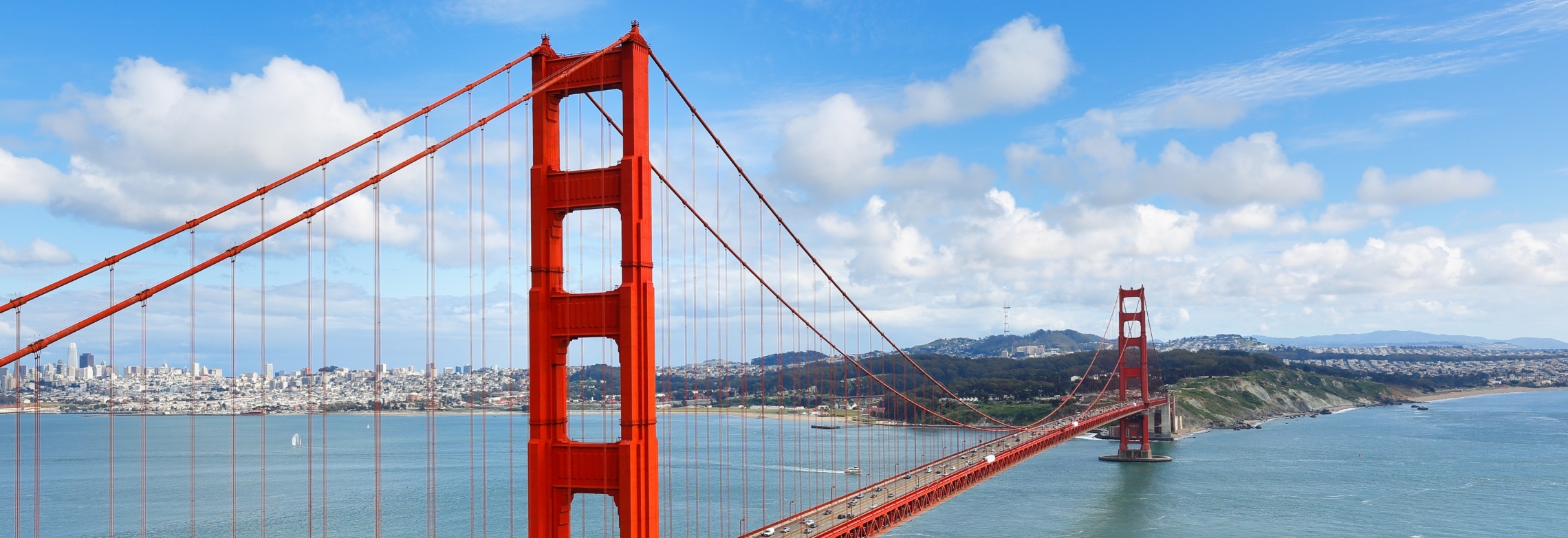 The Golden Gate Bridge stretches across blue water with San Francisco city on the left and green hills on the right, under a partly cloudy sky—an iconic view for travelers flying in after using WallyPark Airport Parking.