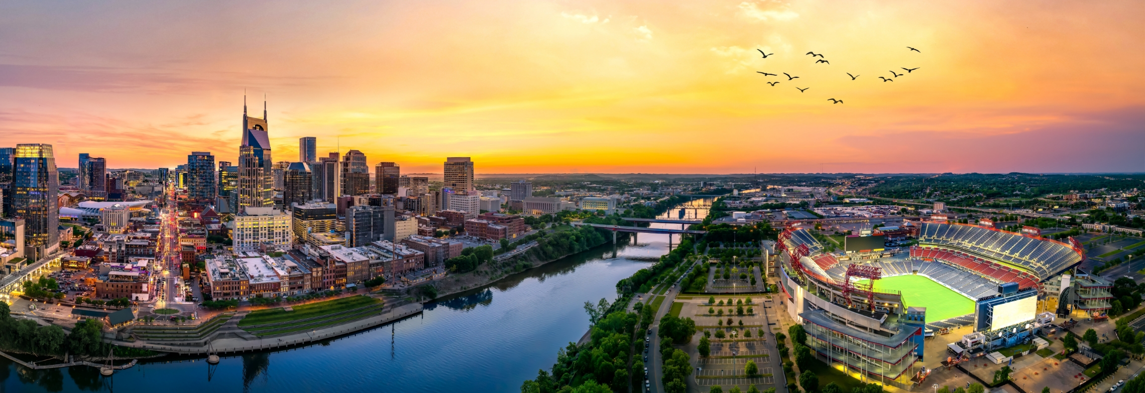 A panoramic view of downtown Nashville at sunset, featuring city skyscrapers, a river, a brightly lit stadium, and a flock of birds flying across a colorful sky—just moments away from WallyPark Airport Parking.