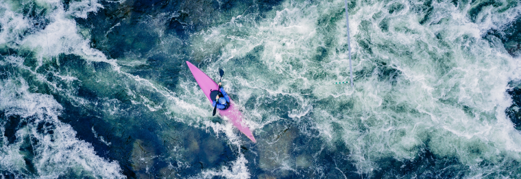 A person in a pink kayak paddles through whitewater rapids, surrounded by churning, foamy water viewed from above—much like navigating the currents at WallyPark Airport Parking during rush hour.
