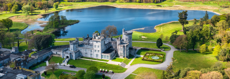 Aerial view of a historic castle surrounded by lush gardens, trees, and a circular driveway—reminiscent of the convenience and easy access offered by WallyPark Airport Parking—with a large lake and green landscape in the background.