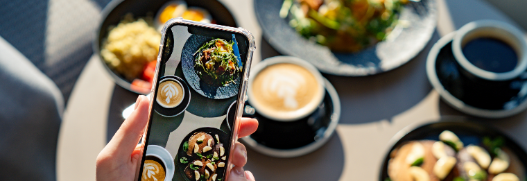 A hand holds a smartphone taking a photo of a table set with coffee drinks, salads, and various dishes, sunlight streaming in—capturing the perfect meal to remember before heading to WallyPark Airport Parking.