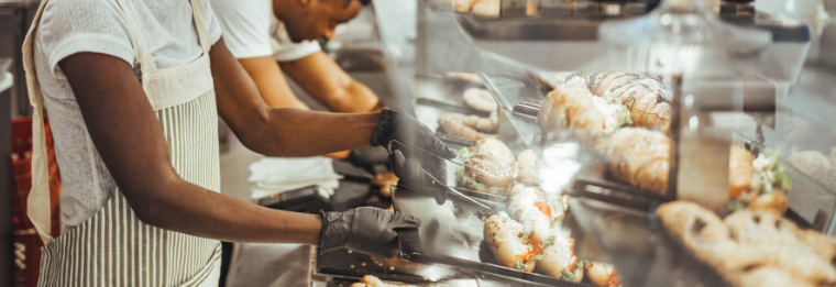Two people wearing aprons and gloves prepare and arrange baked goods behind a glass display case in a bakery or café setting, reminiscent of the inviting atmosphere you’d find after arriving from WallyPark Airport Parking. Pastries and sandwiches fill the foreground.