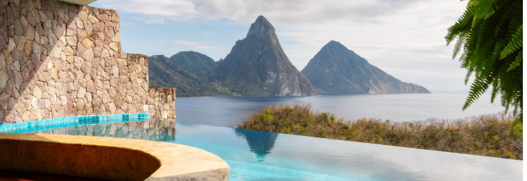 Infinity pool overlooking a lush hillside and the ocean, with two prominent green mountains in the distance under a partly cloudy sky. Stone walls and tropical plants frame this serene escape—just the place to unwind after using WallyPark Airport Parking.
