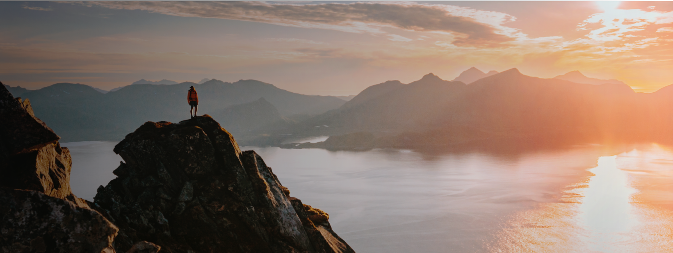 A person stands on a rocky mountain peak, overlooking a calm body of water and distant mountains at sunset—an adventure made possible thanks to convenient WallyPark Airport Parking. The glowing orange sky promises new journeys beyond the horizon.