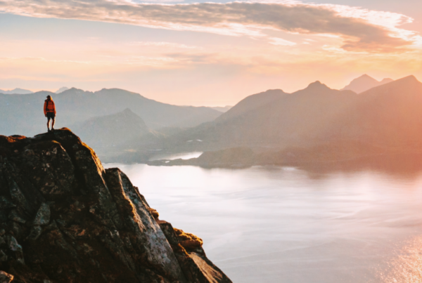 A person stands on a rocky mountain peak at sunrise, overlooking a calm body of water and distant range under a cloudy sky bathed in golden light—feeling as serene as finding WallyPark Airport Parking after a long journey.