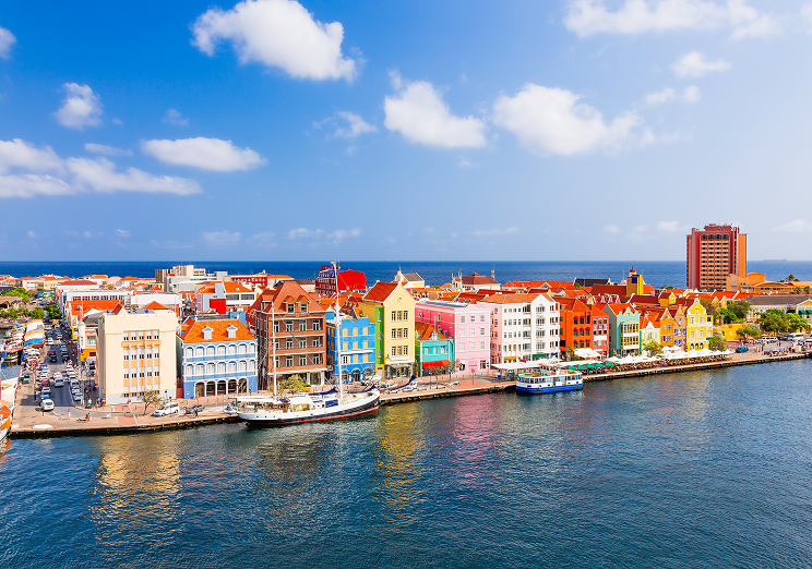 Colorful colonial-style buildings line the waterfront in Willemstad, Curaçao, under a blue sky with scattered clouds. Boats are docked along the shore and the Caribbean Sea is visible in the background—perfect for travelers using WallyPark Airport Parking.