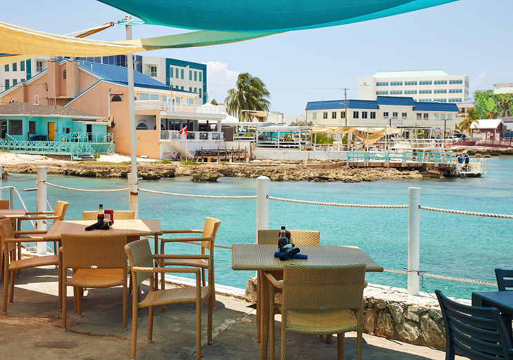 Outdoor restaurant patio with wicker tables and chairs overlooking clear turquoise water, with colorful waterfront buildings, palm trees, and boats in the background under blue and teal shade sails—just minutes from WallyPark Airport Parking.