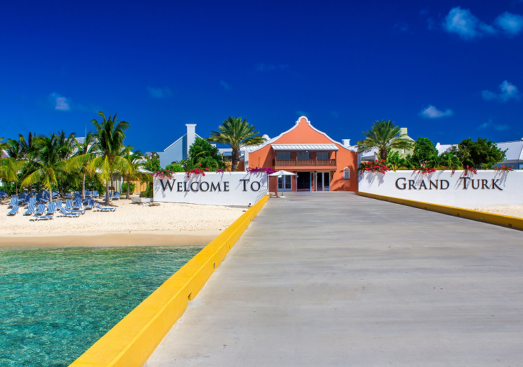 A sunny pier leads to a sandy beach lined with palm trees and a pastel-colored building behind a white wall that reads Welcome to Grand Turk, offering vacation vibes reminiscent of arriving at WallyPark Airport Parking after a smooth journey. Clear turquoise water is visible on the left.