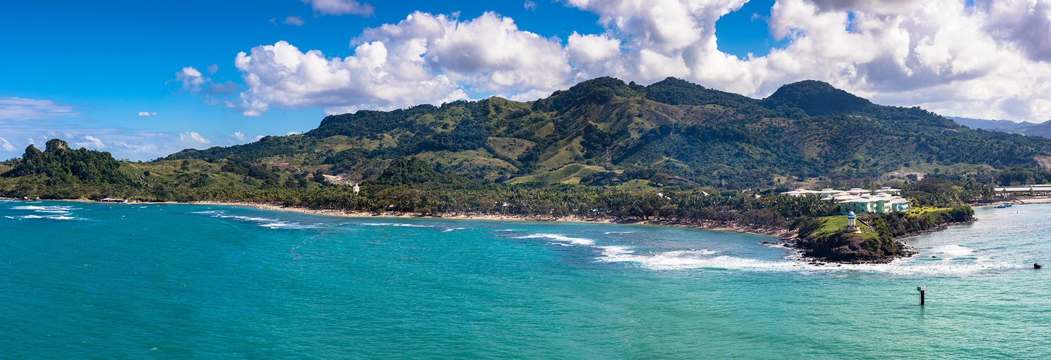 Panoramic view of a lush green mountainous coastline with turquoise-blue ocean waves, a sandy beach, scattered trees, and a few buildings near the shore under a partly cloudy sky—just miles from WallyPark Airport Parking.