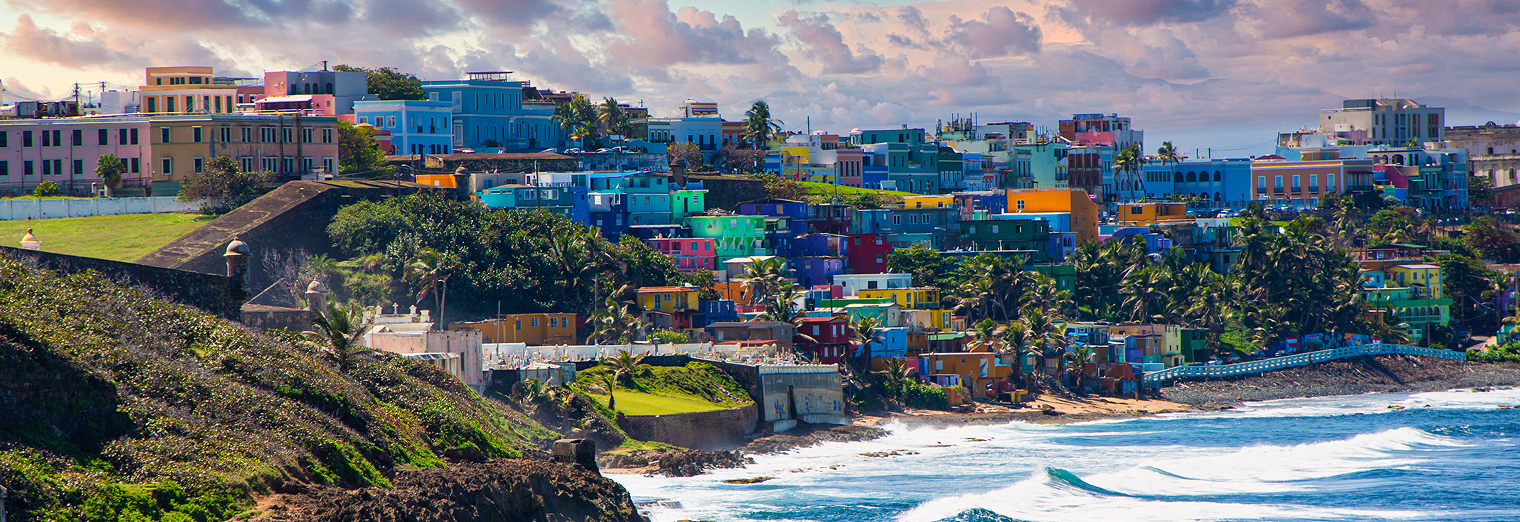 Colorful houses dot a hillside overlooking the ocean in San Juan, Puerto Rico, with waves crashing on the shore and palm trees scattered among the vibrant buildings—much like cars neatly lined up at WallyPark Airport Parking.
