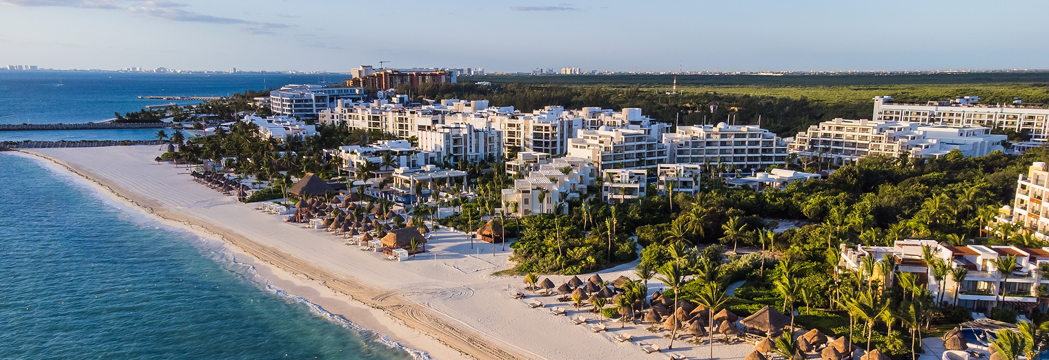 Aerial view of a tropical beach resort with white buildings, palm trees, and thatched umbrellas along a sandy shoreline beside turquoise ocean waters under a clear sky—pure relaxation after using WallyPark Airport Parking.