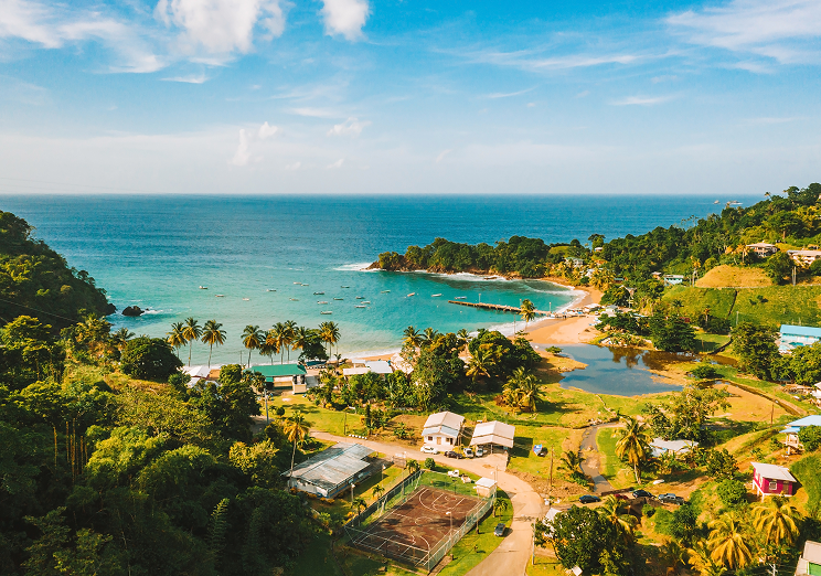 Aerial view of a tropical coastline with turquoise water, sandy beaches, palm trees, lush greenery, small buildings, and a few boats along the shore under a bright blue sky—ideal for dreaming of travel after using WallyPark Airport Parking.