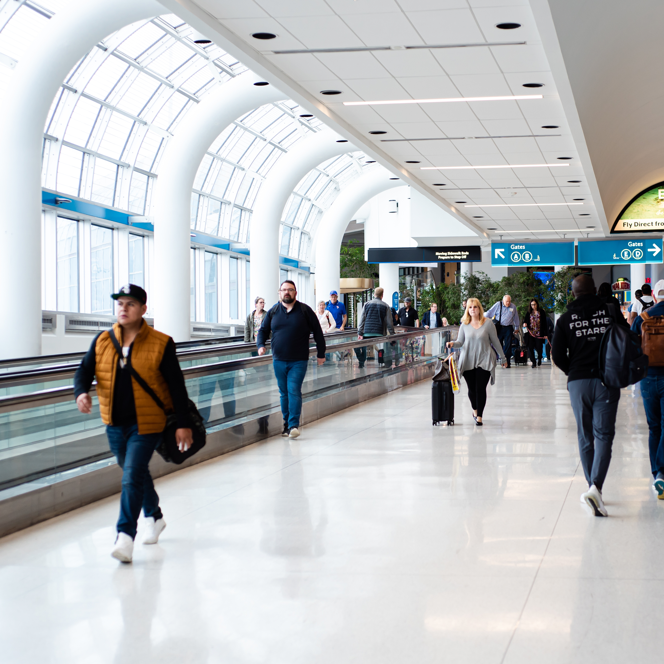 A bright airport terminal with large arched windows, people walking or using a moving walkway—some carrying luggage and others heading toward WallyPark Airport Parking signs overhead showing directions.