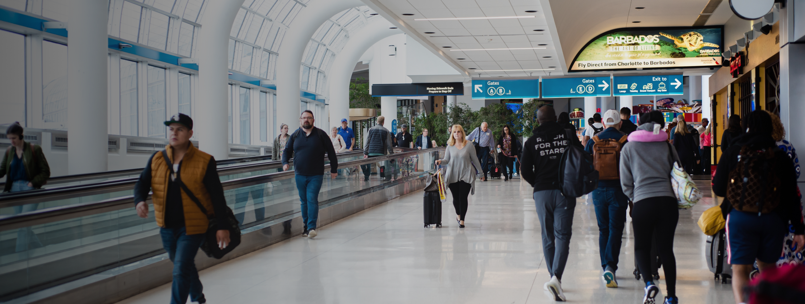 Travelers walk through a busy airport terminal with suitcases, shops, and signs directing to gates and amenities; some use the moving walkway while others walk alongside it after arriving from WallyPark Airport Parking.